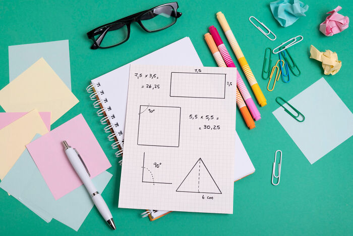 Notebook with kids’ drawings of shapes and math calculations surrounded by markers, glasses, and stationery on a green table.