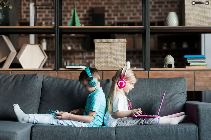 Two kids wearing headphones, sitting back to back on a couch playing with laptop and gaming device, childhood memories concept.