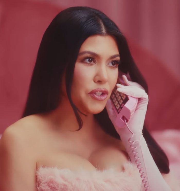 Kourtney Kardashian holding pink phone, promoting lollipops for women&rsquo;s private parts, sparking reactions in a pink setting.