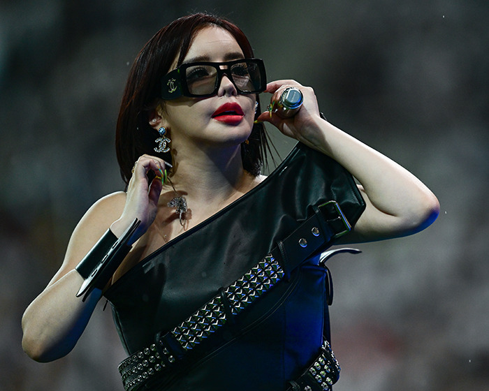 K-Pop icon Park Bom wearing black leather outfit and oversized sunglasses during a dramatic plastic surgery transformation reveal.