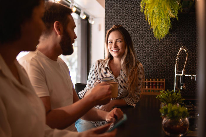 Woman nervously smiling while talking to an elite military diver in a cozy cafe, holding a coffee cup during their date.