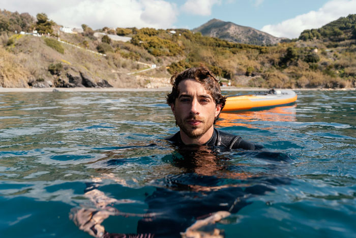 Man in wetsuit floating in the sea near a paddleboard with coastal hills in the background, elite military diver concept.