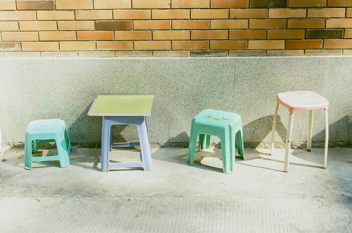 A row of colorful plastic stools and a small table placed outside against a sunlit brick and concrete wall.