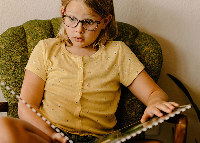 Young girl with glasses sitting on a green chair, reading a book with a surprised expression about steak and cows.