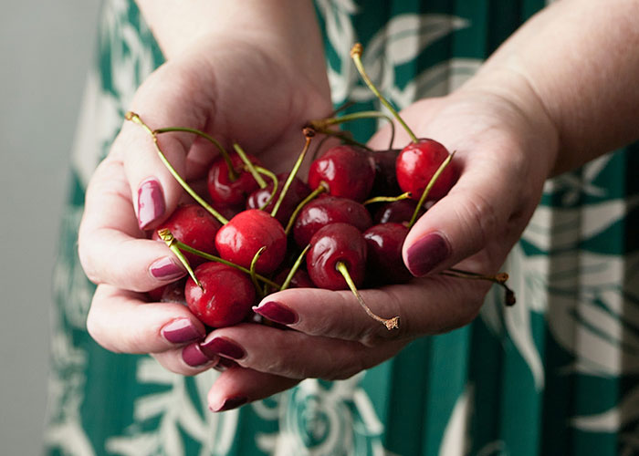 Hands holding a bunch of fresh red cherries with stems, against a blurred patterned green dress background.