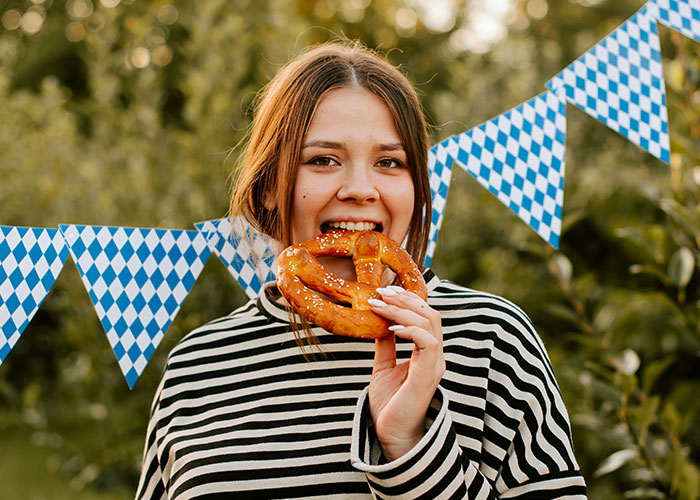 Young woman in striped shirt eating a pretzel outdoors with blue and white flags in the background about cows and steak facts.