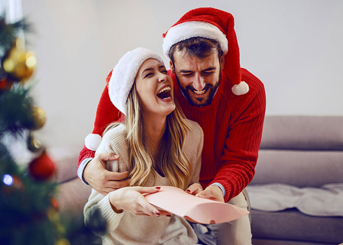 Couple wearing Santa hats laughing and sharing a Christmas gift, enjoying a joyful moment during the holiday season.