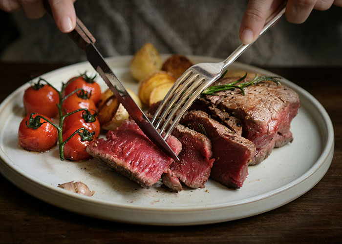 Close-up of a person cutting rare cooked steak on a plate with tomatoes and roasted potatoes, illustrating steak from cows.