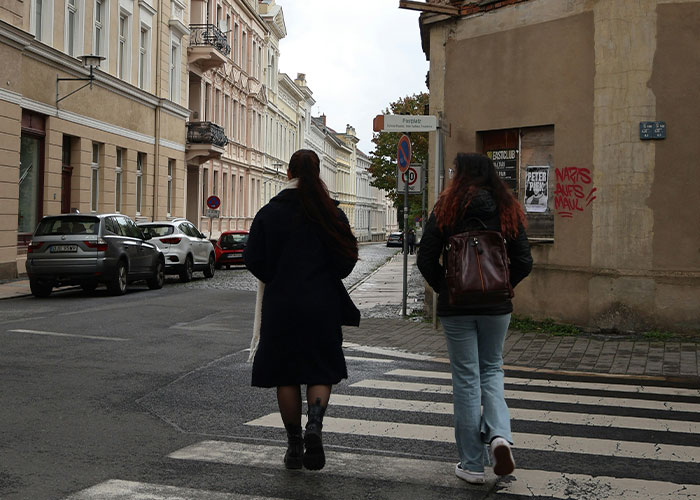 Two women walking on a city crosswalk near parked cars and buildings, illustrating everyday life scenes.