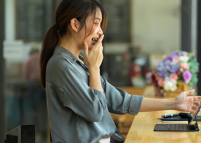 Young woman yawning while using a laptop in a casual workspace, illustrating the concept of learning facts about steak and cows.