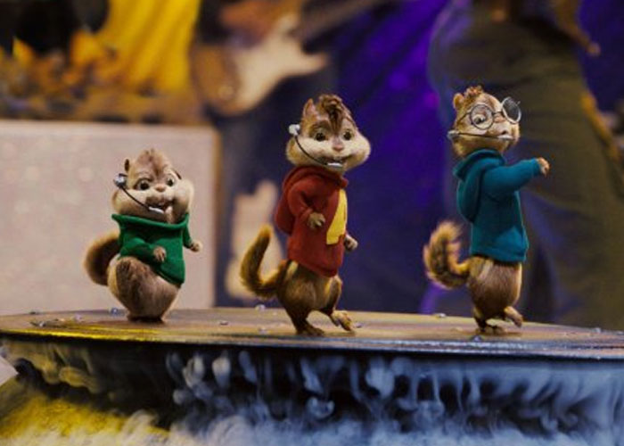 Three animated chipmunks wearing colorful hoodies performing on stage with musical instruments in the background.