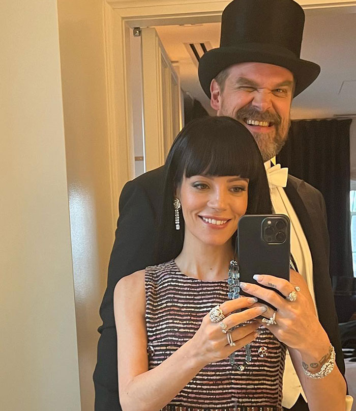 David Harbour dressed in a tuxedo and top hat stands behind a smiling woman taking a selfie with a smartphone.