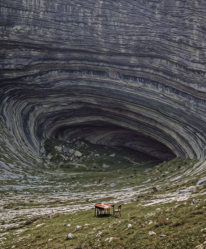 A solitary table and chair set in a vast, unnerving liminal space with layered rock formations and grass.