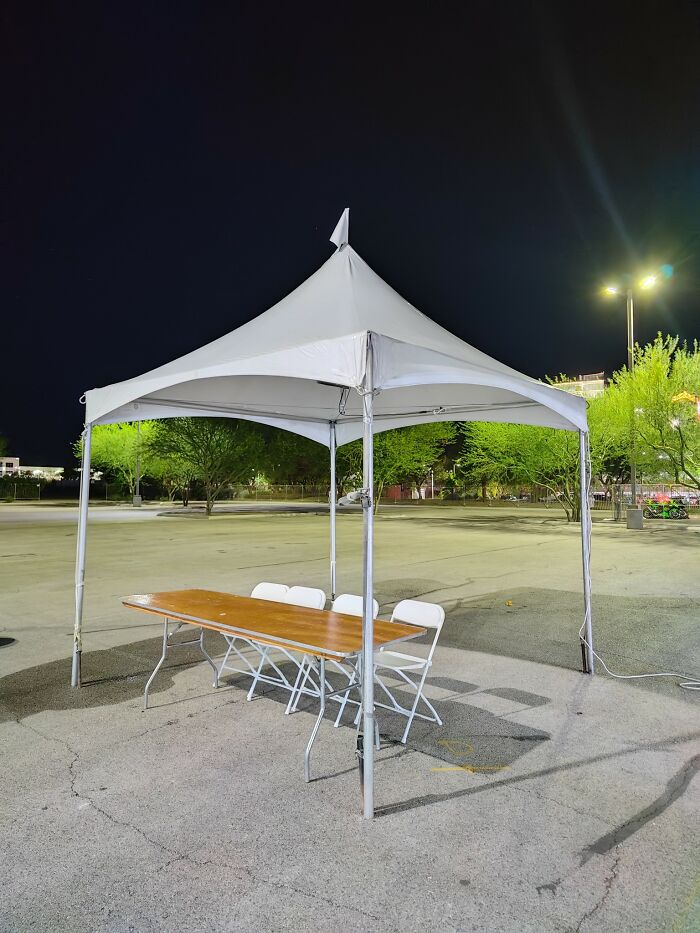 Empty outdoor tent with table and chairs in a quiet, liminal space at night, evoking a surreal and unnerving atmosphere.