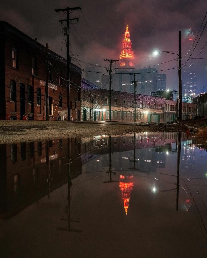 Urban liminal space at night with brick buildings, power lines, and illuminated tower reflections in a puddle.