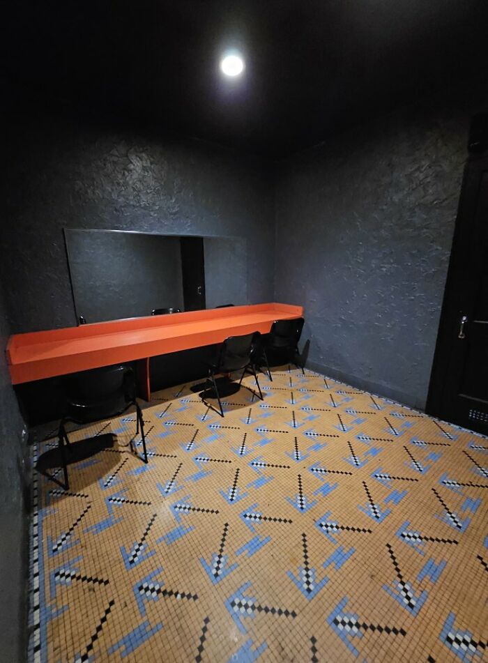 Dimly lit room with textured walls, bright orange counter, black chairs, and patterned tile floor, a liminal space with eerie vibe.