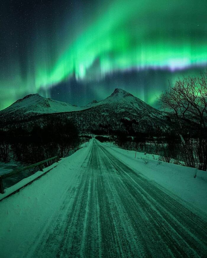 Snow-covered road under the northern lights with dark mountains and trees creating a beautiful yet unnerving liminal space.