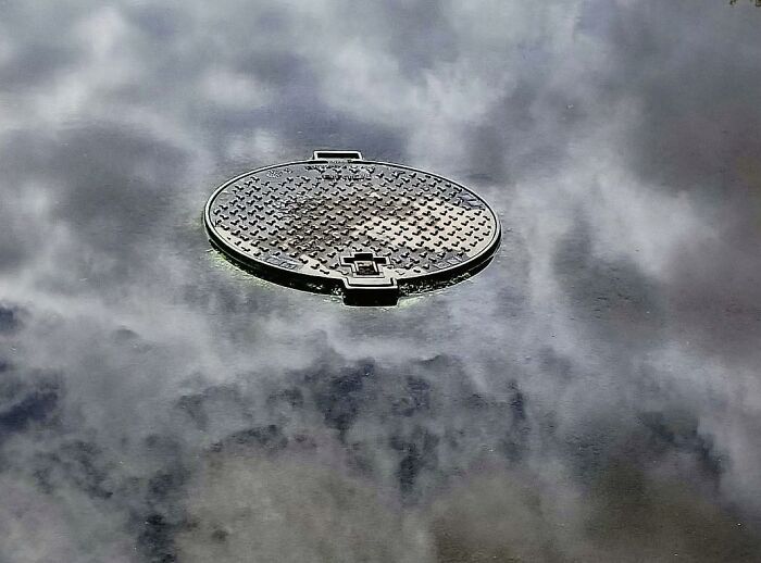 Circular manhole cover reflecting cloudy sky, creating a beautiful yet unnerving liminal space scene.