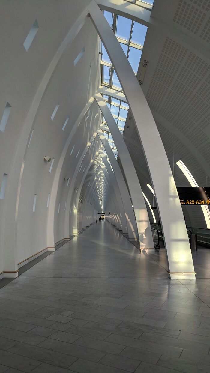 Long, empty architectural hallway with tall white arches and skylights creating a surreal liminal space atmosphere.