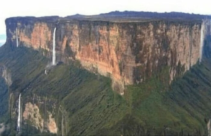 Massive flat-topped mountain with waterfalls flowing down steep cliffs in a beautiful yet unnerving liminal space.