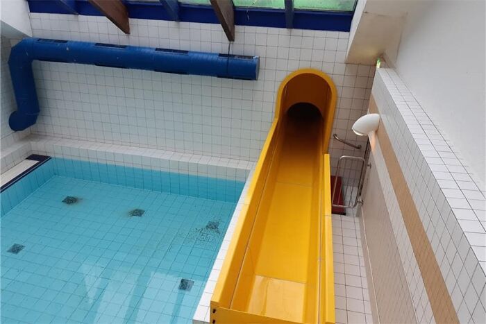 Empty indoor pool with a yellow slide, showcasing a beautiful yet unnerving liminal space with bright tiles and exposed beams.