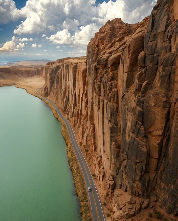 A narrow road running alongside a turquoise river with towering red cliffs, creating a beautiful yet unnerving liminal space.