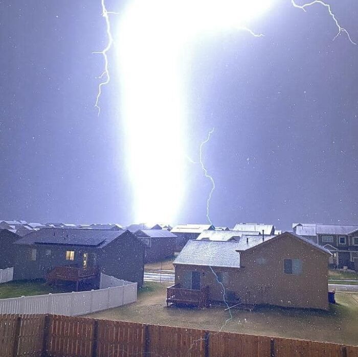 Bright lightning striking near suburban houses, creating an unnerving liminal space with intense light and shadow contrasts.