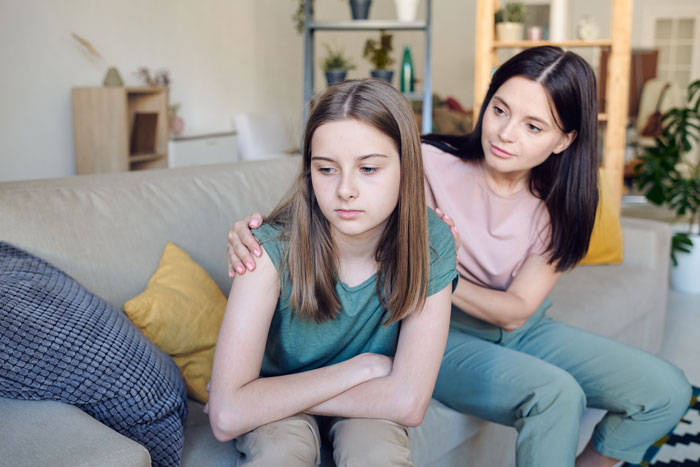 Teen girl looks upset on couch as woman comforts her, depicting family tension and stepsister setting boundaries.