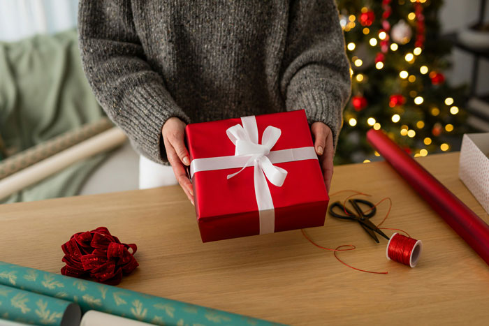 Person holding red gift box with white ribbon near Christmas tree, illustrating stepsister boundaries and gift conflict.