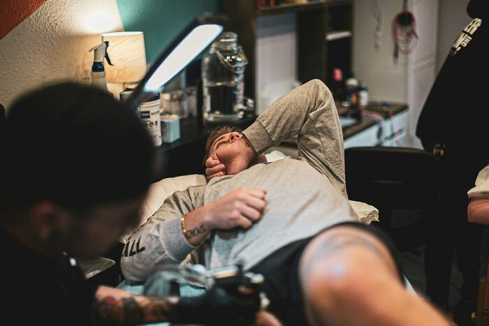 Man lying on a table covering his face while tattoo artist works on his leg in a tattoo studio setting.