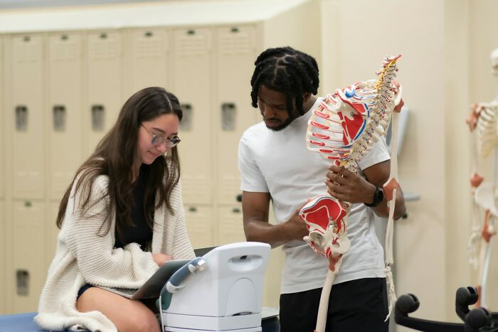 Two medical students studying anatomy using a skeletal model in a classroom setting with lockers in the background