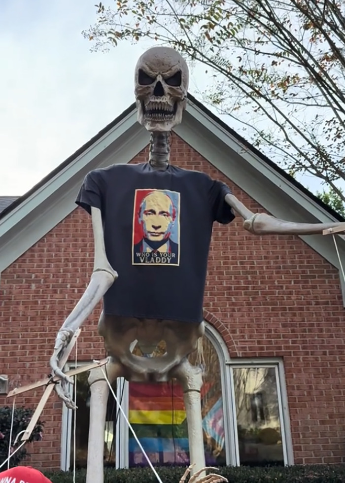 Skeleton Halloween display wearing a political shirt in an elaborate anti-MAGA decoration with colorful flags outside a brick house.