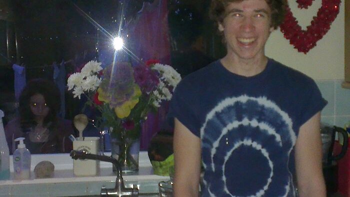 Young man smiling in a kitchen with a creepy doll-like figure and glowing eyes reflected in the dark window behind him.