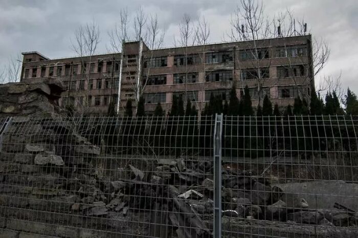Abandoned building behind a damaged fence and leafless trees, evoking a creepy photo moment.