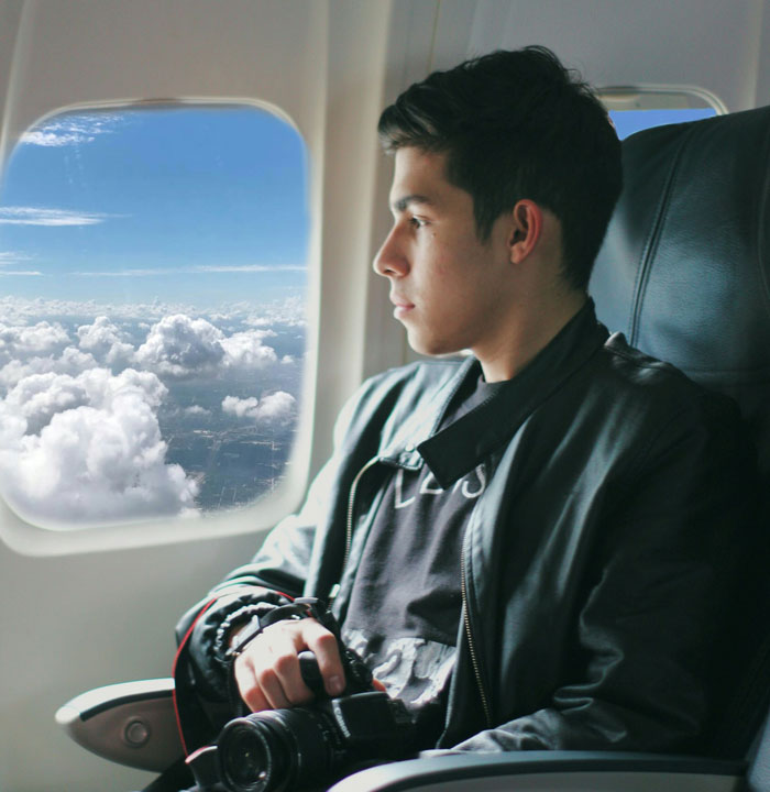 Teen refusing to entertain a stranger&rsquo;s kid on a flight, sitting by the window holding a camera, looking outside.