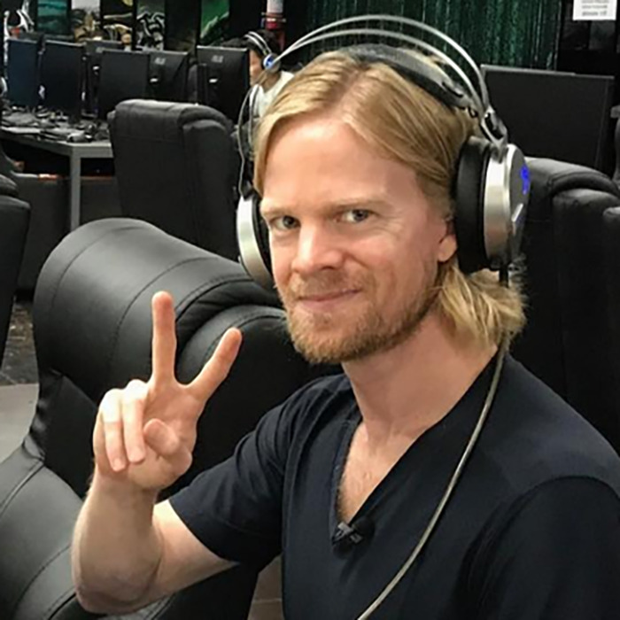 Man behind blinking guy GIF wearing headphones and black shirt, making a peace sign indoors with computer setups in the background.