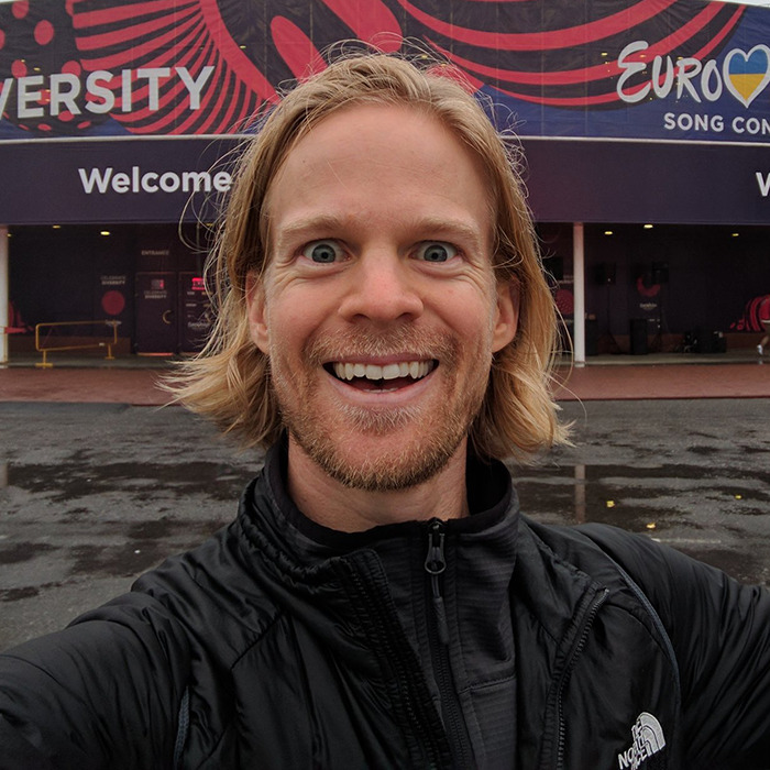 Man behind blinking guy GIF selfie with long hair and beard, smiling outside a building with colorful banners.