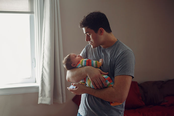 Father holding and looking at baby in a dimly lit room, illustrating burnt-out wife asking husband to feed the baby. Father holding and looking at baby in a dimly lit room, illustrating burnt-out wife asking husband to feed the baby.
