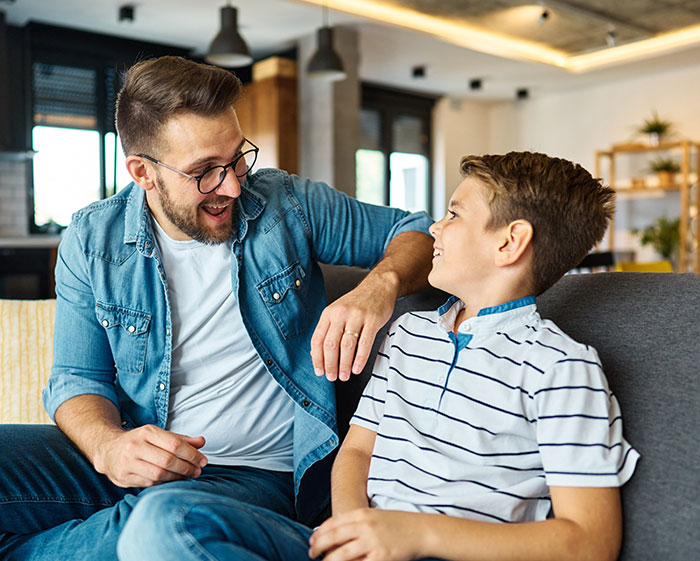 Man and boy sitting on couch smiling at each other, reflecting a father and son relationship with uncertainty about real parents.