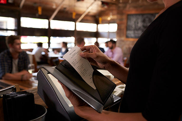 Person holding a bill inside a restaurant with guests in the background after entitled guests go crazy ordering