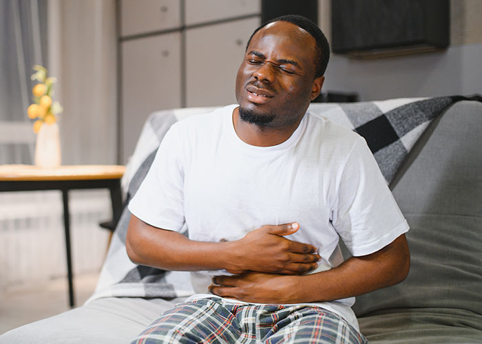 Man in casual clothes sitting on couch, clutching stomach in discomfort, showing true colors during vacation.
