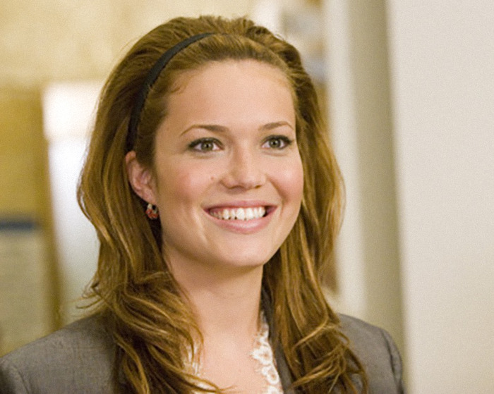 Mandy Moore smiling with long hair and a gray blazer in a softly lit indoor setting.