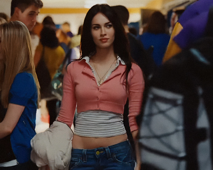 Megan Fox wearing a pink cropped top and jeans, sparking plastic surgery speculation at a crowded indoor event.