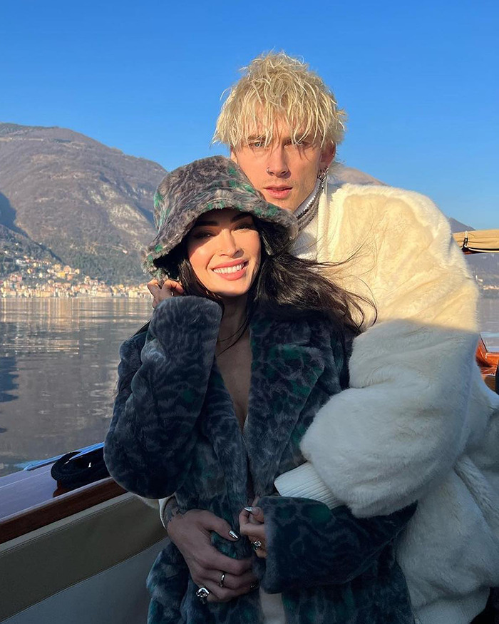 Megan Fox wearing a patterned coat and hat, smiling and posing outdoors with a man in a white fur jacket by the lake.