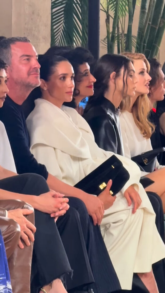 Meghan Markle sitting front row at Paris Fashion Week, dressed in white, holding a black clutch amid other fashion attendees.