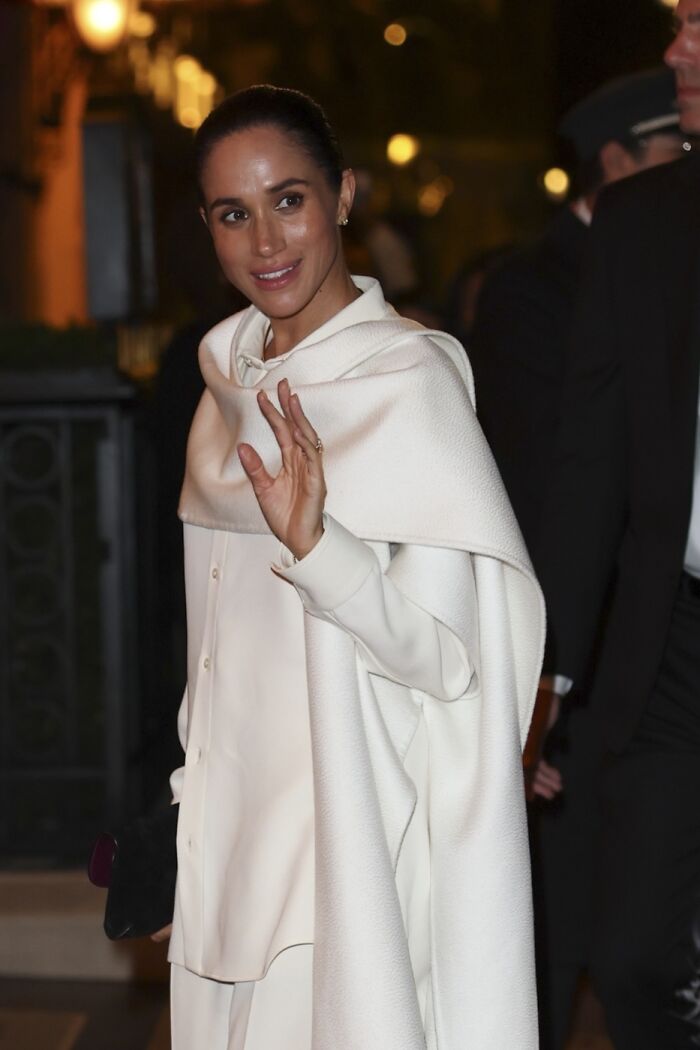 Meghan Markle smiling and waving at Paris Fashion Week, wearing an elegant white outfit during a nighttime event.
