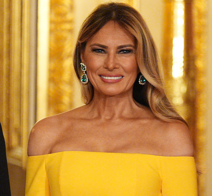 Melania Trump wearing a yellow off-shoulder dress and emerald earrings at a White House event with golden decor background