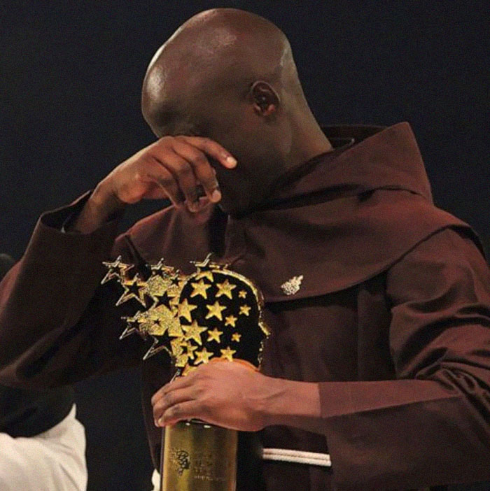 A man in a brown robe showing emotion while holding a golden trophy with star-shaped decorations, men are good.