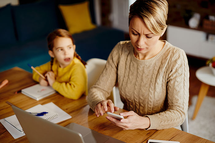 Mother working on smartphone at table while daughter in yellow sweater looks on, highlighting toxic in-laws conflict and newborn issues.