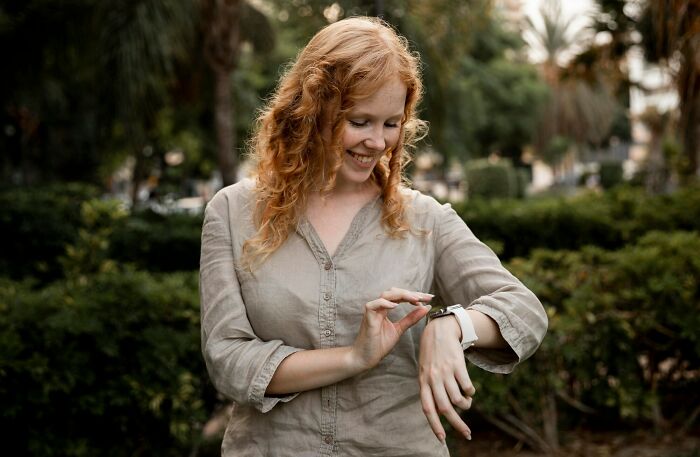 Red-haired woman outdoors smiling while looking at her smartwatch, capturing moments of misreading social cues.